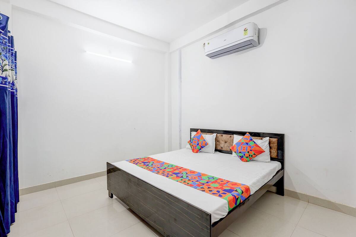 Una cama individual con estructura de madera, ropa de cama blanca y almohadas con estampados geométricos coloridos. Un equipo de aire acondicionado está montado en la pared sobre la cama.