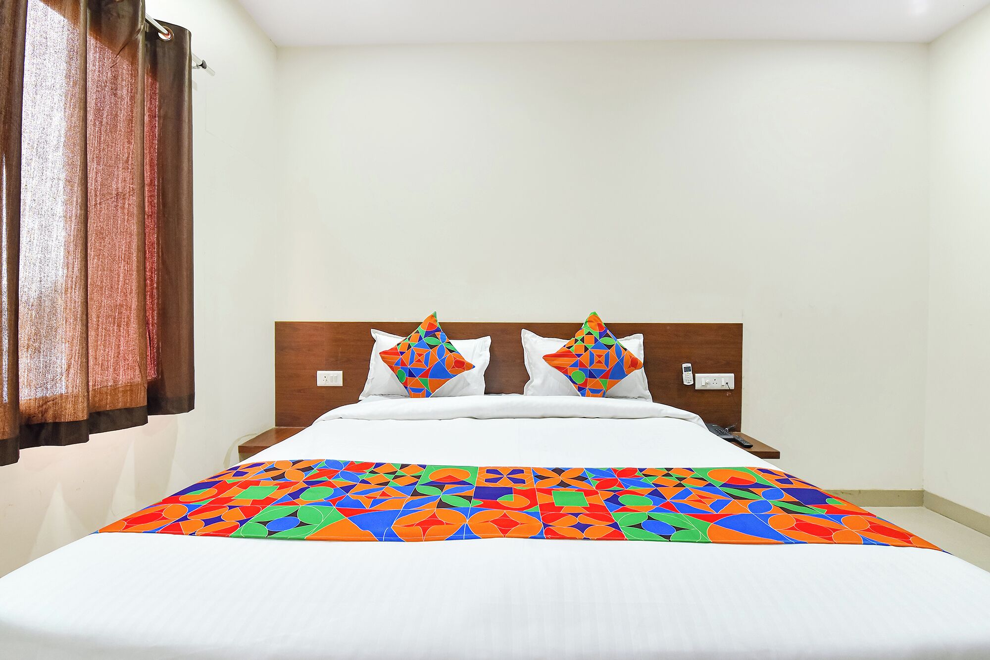 A hotel room with a bed, a wooden headboard, two pillows, a white bedspread, and a window with curtains.