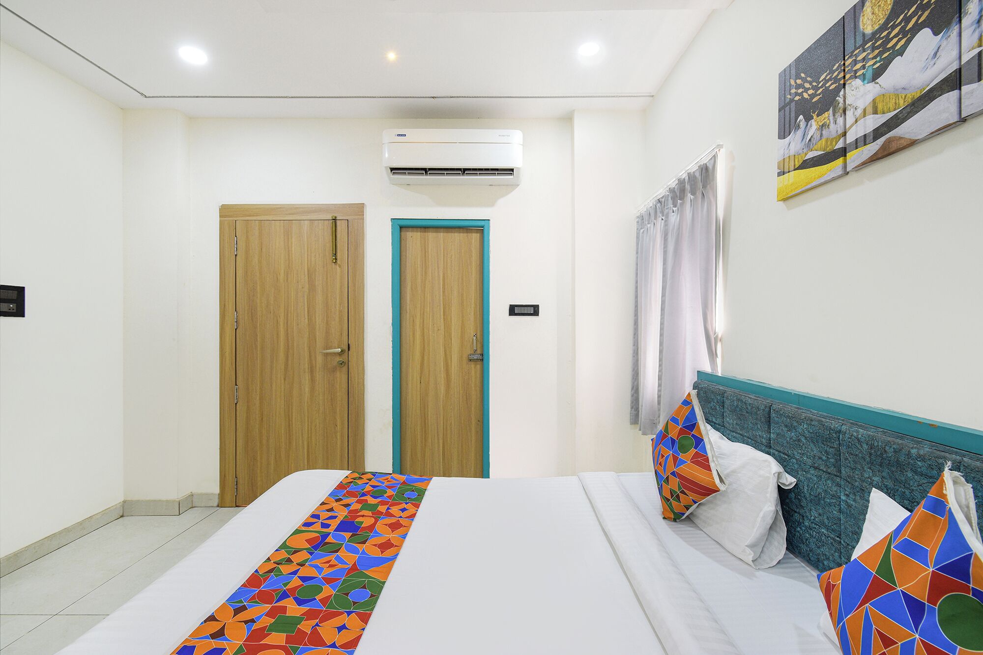 A hotel room with a bed, a wooden door, an air conditioning unit, a wall-mounted light, and a painting.