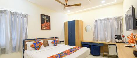 A hotel room with a bed, a desk, a wardrobe, a ceiling fan, a painting, and a television.