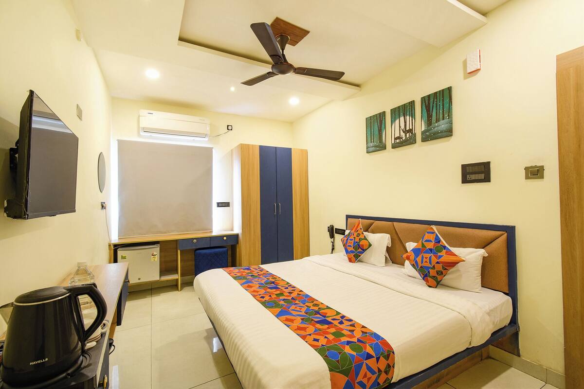 Quarto de hotel com cama, escrivaninha, cafeteira, ventoinha no teto e obras de arte na parede.