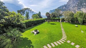 A well-maintained garden with a clear path leading to a seating area, surrounded by lush greenery and a view of a mountainous coastline. - Nice apartment for 4 guests with A/C, WIFI, TV and patio (Riva Del Garda)