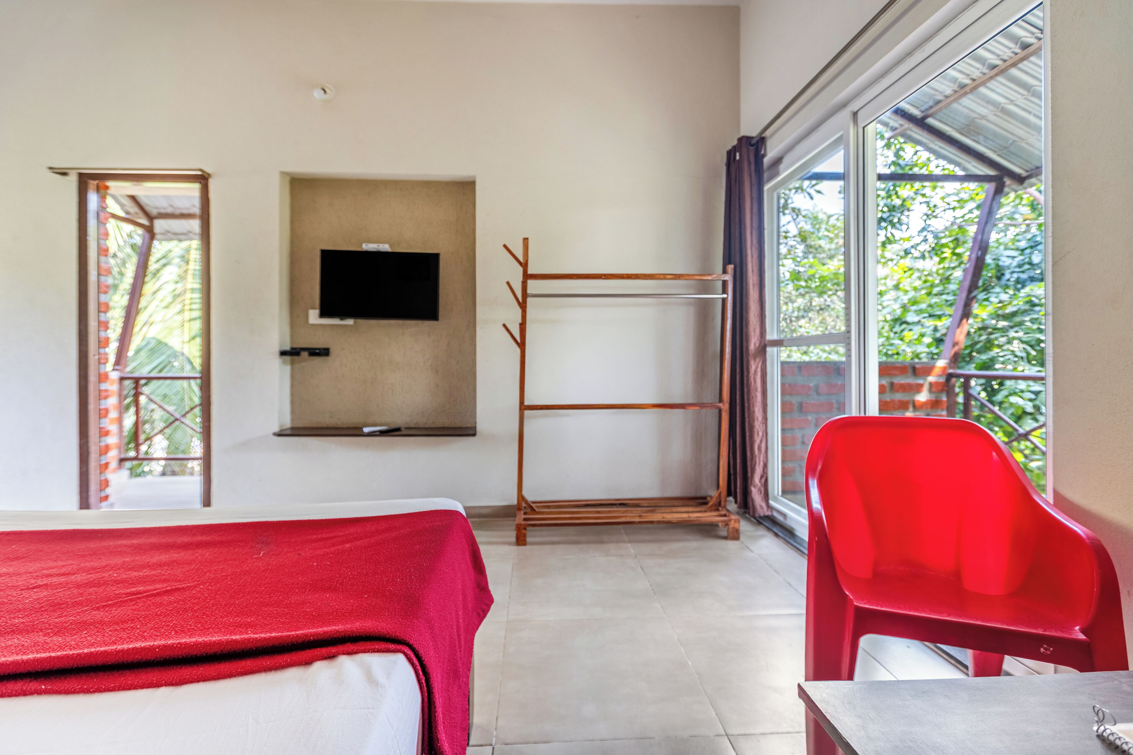 A room with a bed, a TV mounted on the wall, a drying rack, a red chair, and a window with a view of greenery.