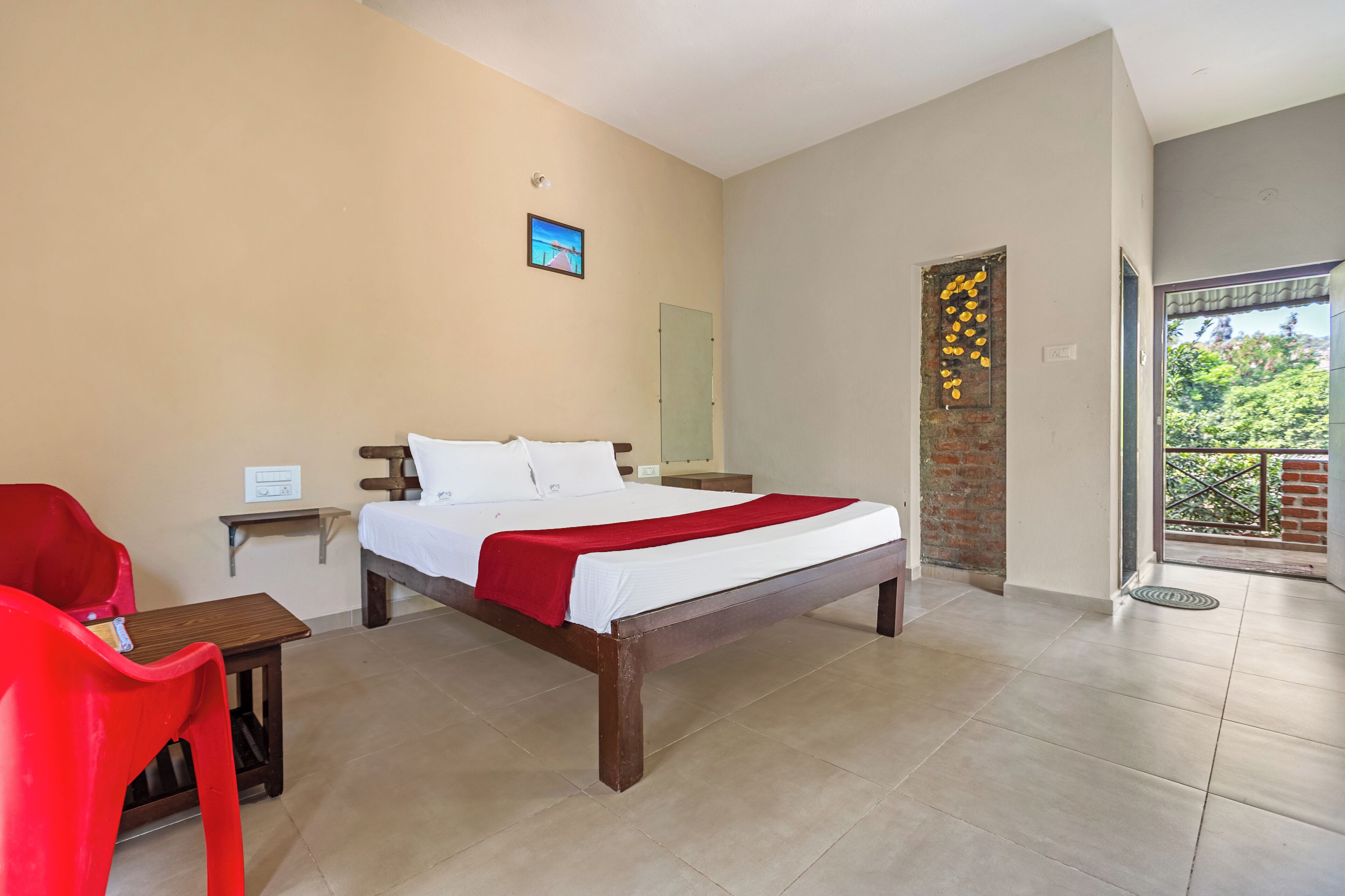 A bedroom with a bed, a red chair, a small table, a nightstand, a door with a decorative panel, and a view of greenery through a glass door.