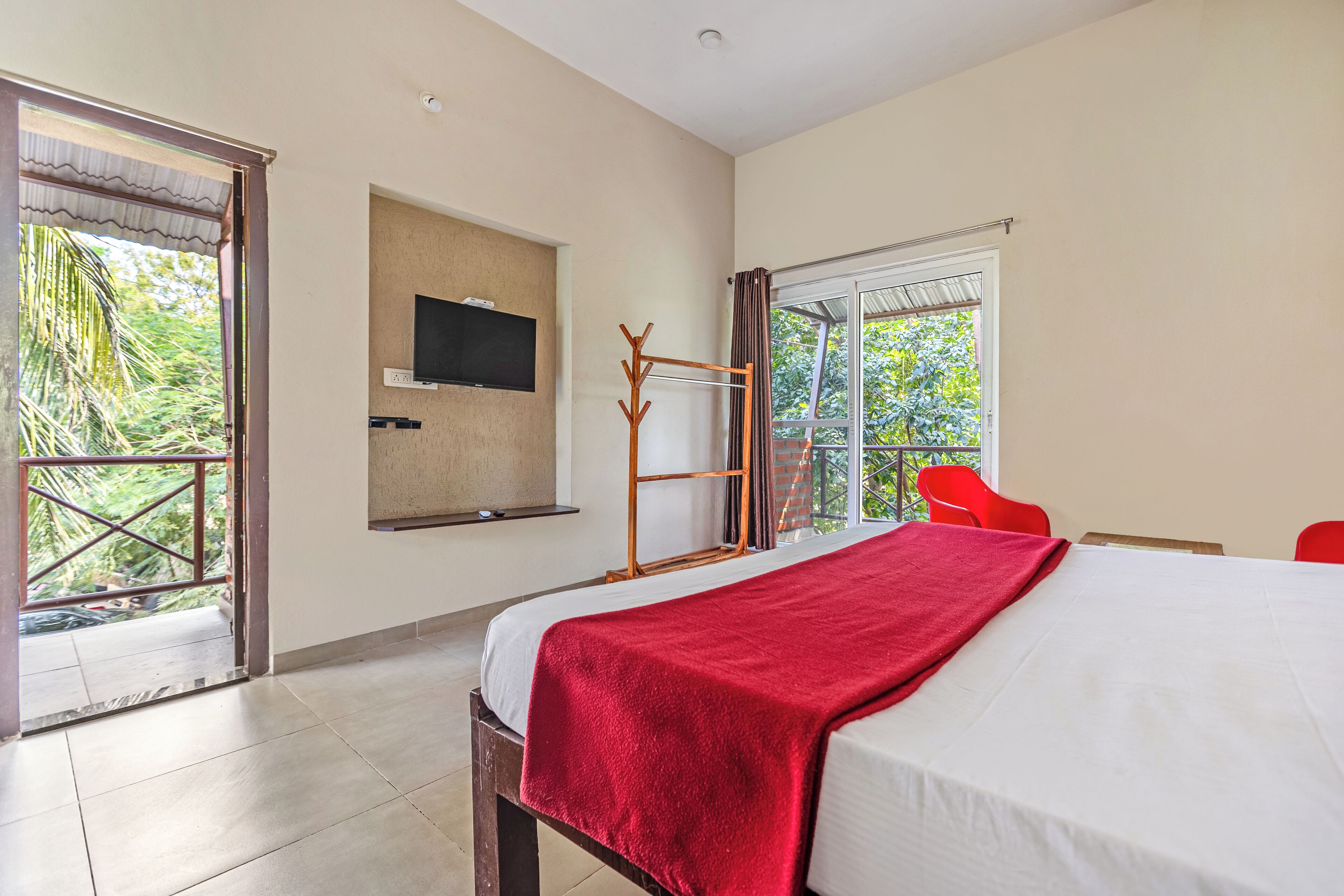 A hotel room with a bed, a TV mounted on the wall, a balcony with a view of greenery, and a red chair.