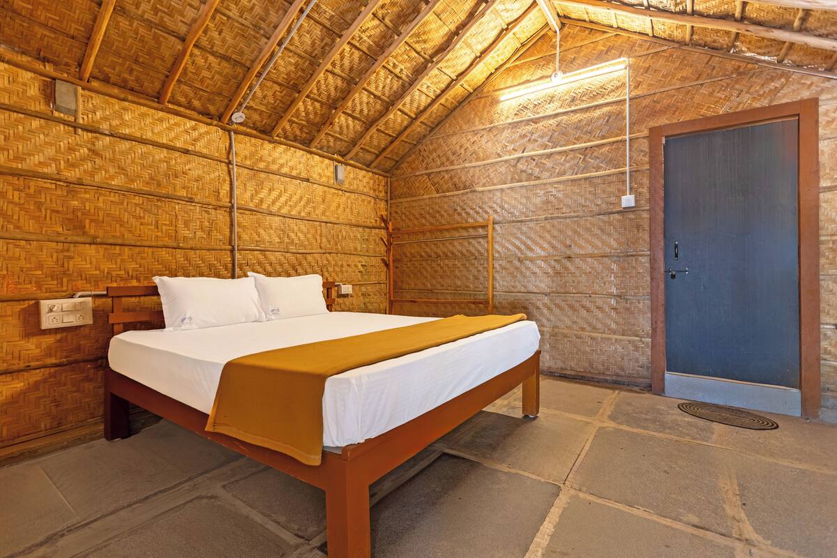Una cama individual con estructura de madera y ropa de cama blanca, una puerta a la derecha y un techo de paja.