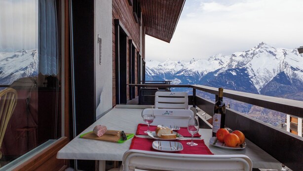 Ein Esstisch mit Blick auf schneebedeckte Berge, eine Weinflasche und ein Teller Essen auf einem Balkon.