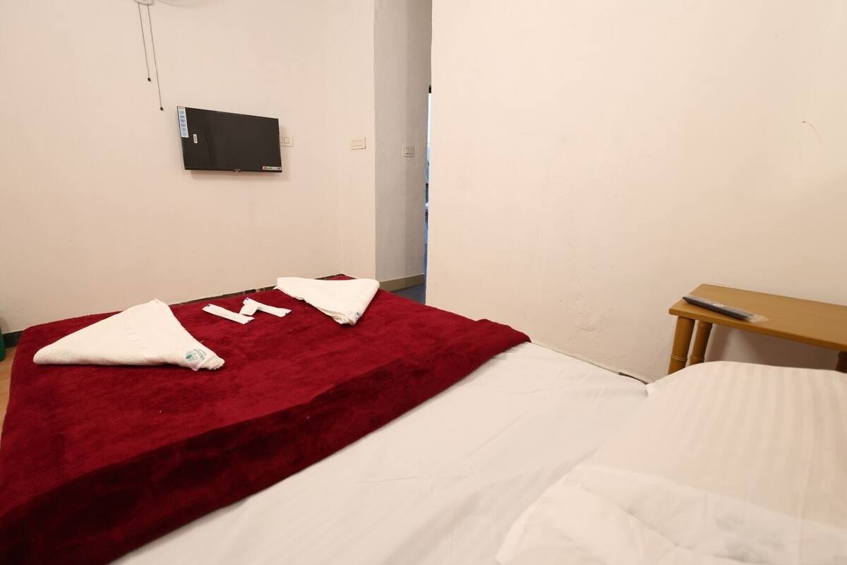 Ein Einzelbett mit roter Decke und weißen Laken, ein Nachttisch aus Holz sowie ein flachbildfernseher an der Wand.