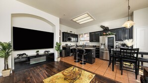 A modern kitchen with dark cabinetry, a large island, and a dining area with black chairs. - Luxurious 5 Bedroom Retreat with Saltwater Pool (Elk Grove)