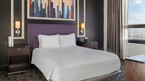 Habitación de hotel con una cama grande, un cuadro de paisaje urbano, dos mesitas de noche con lámparas y una ventana con cortinas.