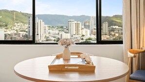 A round wooden table with a white vase and a tray containing a decorative item, positioned by a window with a cityscape view. - Waikiki Island Skyline Serenity with Mountain Views (Honolulu)