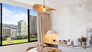 A modern room with a round wooden table, two chairs, a hanging light fixture, a desk with a lamp, and a view of buildings through large windows. - Waikiki Island Skyline Serenity with Mountain Views (Honolulu)