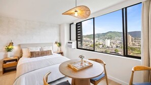 A modern hotel room with a large bed, a dining table, and a city view through large windows. - Waikiki Island Skyline Serenity with Mountain Views (Honolulu)