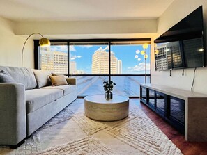 A modern living room with a sofa, a round coffee table, a TV mounted on the wall, and a large window with a city view. - Chic Downtown Honolulu Retreat – Free Parking (Honolulu)