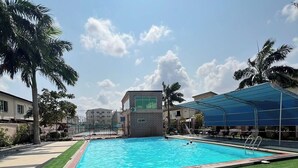 A swimming pool with a tiled deck, surrounded by palm trees and a covered area with chairs. - Recks Luxury Homes (Abuja)
