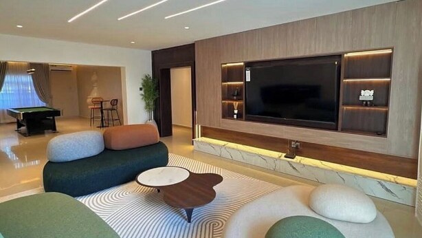 A modern living room with a sofa, ottomans, a coffee table, and a TV mounted on the wall. - Recks Luxury Homes (Abuja)