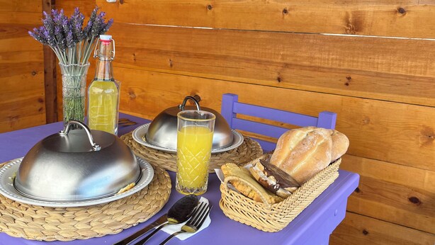 A table with a covered silver dish, a glass of orange juice, a basket of bread, and a bottle of olive oil. - Natural Romance in Glamping Rincón de Lavanda (Texcoco de Mora)