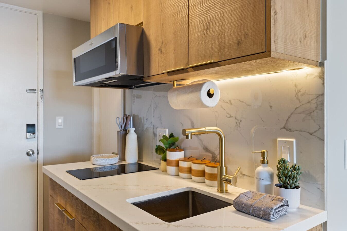 A modern kitchen with wooden cabinets, a stainless steel microwave, a roll of paper towels, a sink with a gold faucet, and jars of spices.
