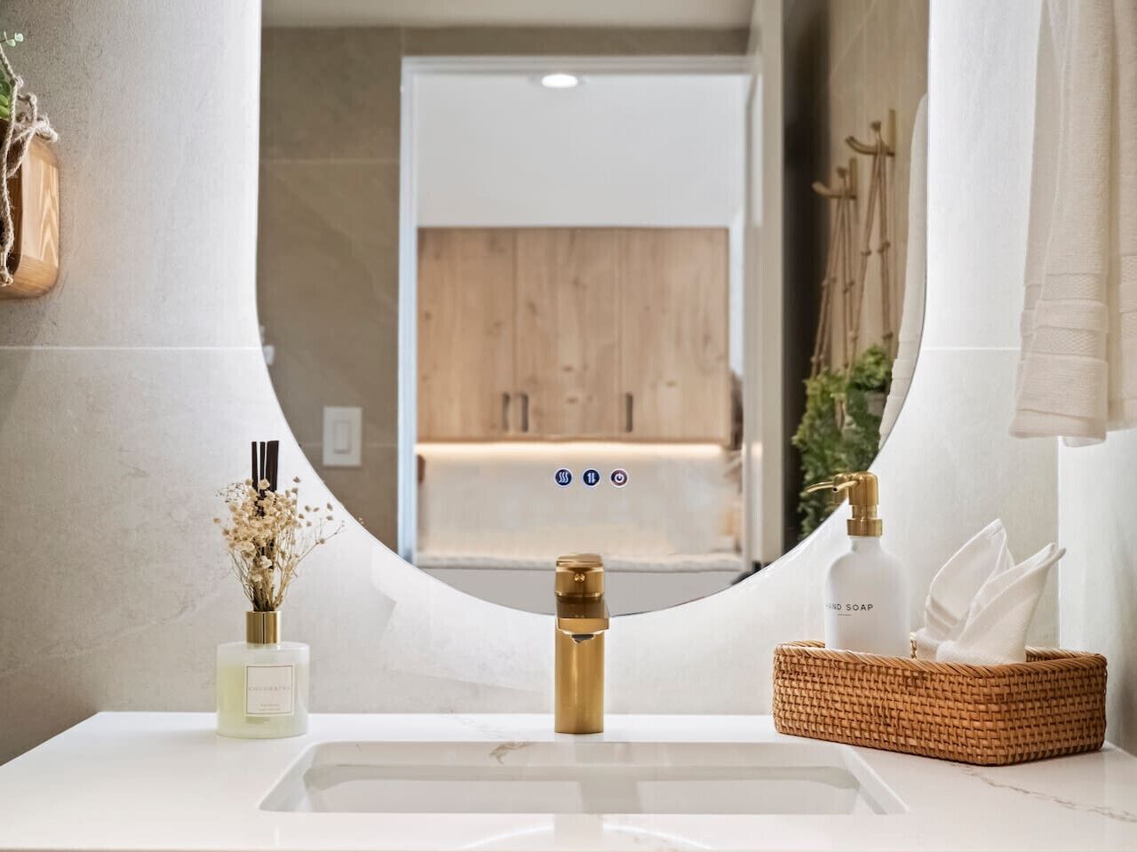 A bathroom with a white sink, a gold faucet, a bottle of soap, a vase with dried flowers, and a woven basket with a towel.