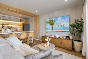 A modern living room with a sofa, coffee table, and a dining area with chairs. There's a large TV mounted on the wall and a potted plant. - Waikiki Luxury Ocean Escape 33F – 1 Parking (Honolulu)