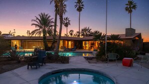 Una piscina con zona de patio, palmeras y una casa con luz cálida.