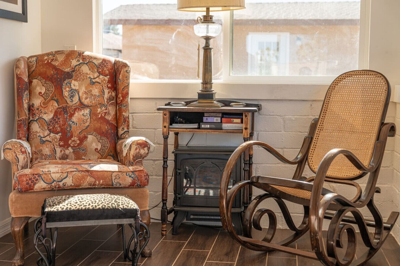 A room with a patterned armchair, a wicker rocking chair, a small table with a lamp, and a fireplace.