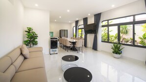 Een moderne woonkamer met een bank, twee ronde tafels, een eethoek met stoelen en grote ramen met planten.