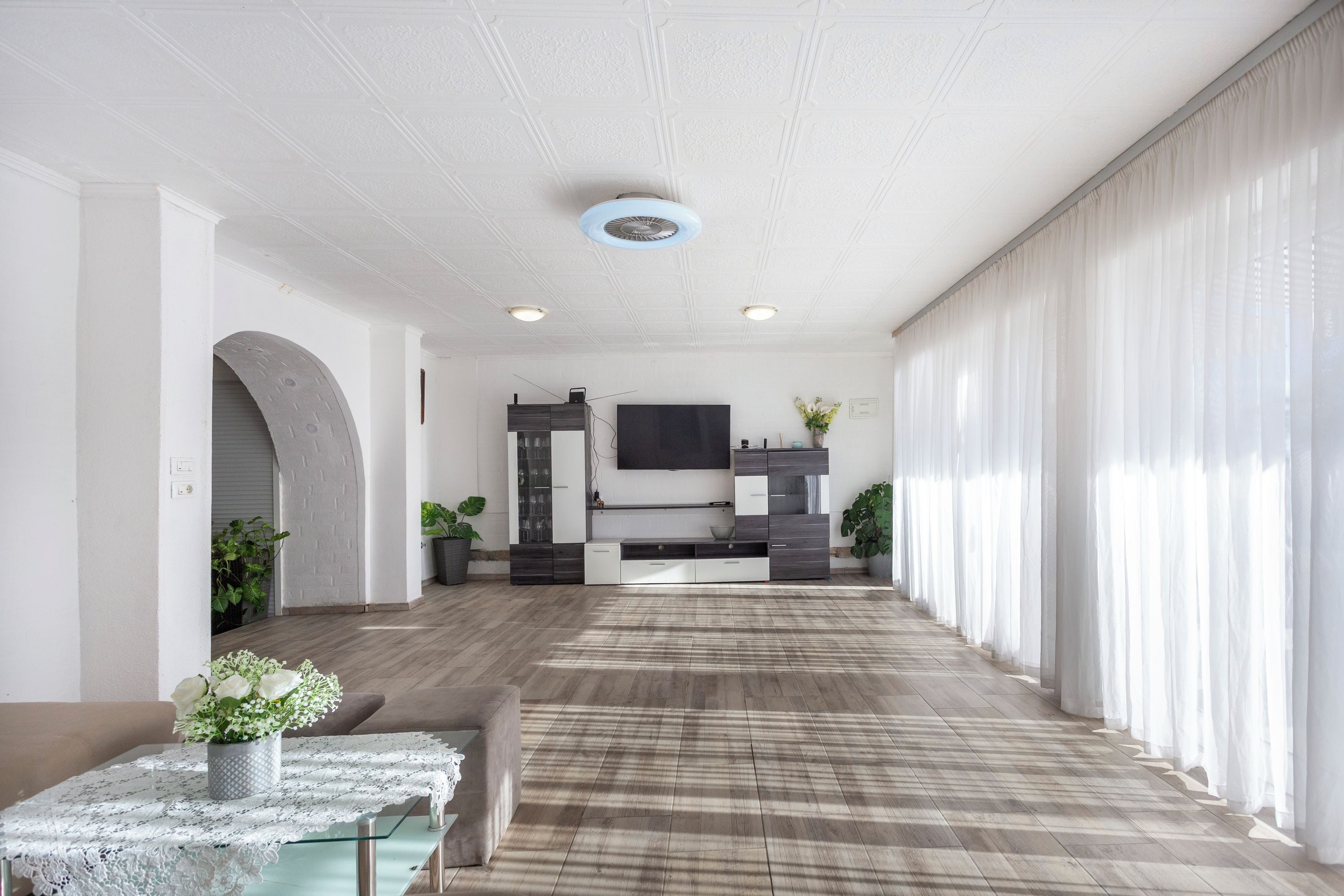 A spacious living area with a wooden floor, a glass coffee table with a lace tablecloth, a flat-screen TV mounted on the wall, and a large window with sheer curtains.