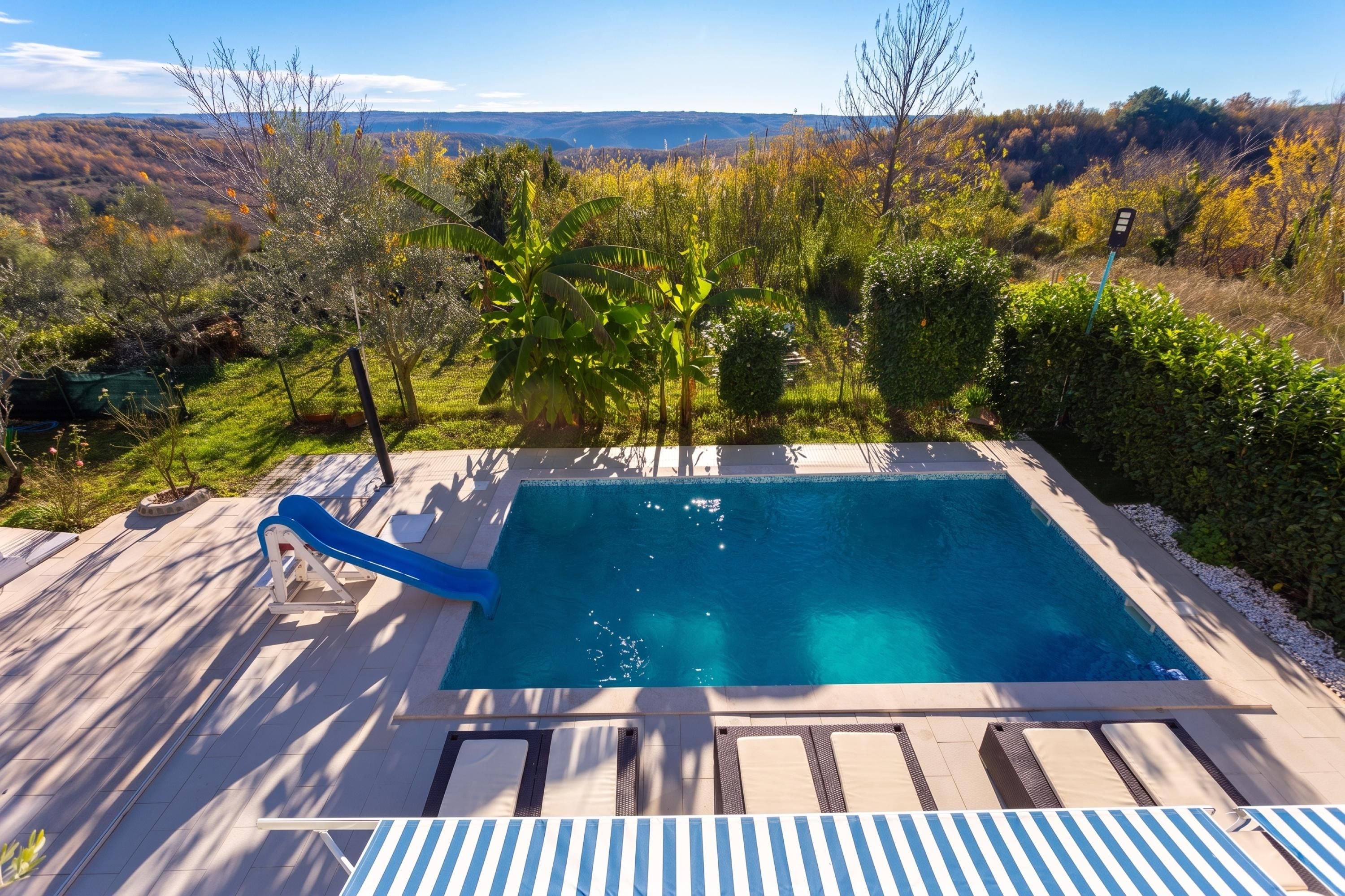 A rectangular swimming pool with a slide, surrounded by a deck with lounge chairs and a view of a hilly landscape.