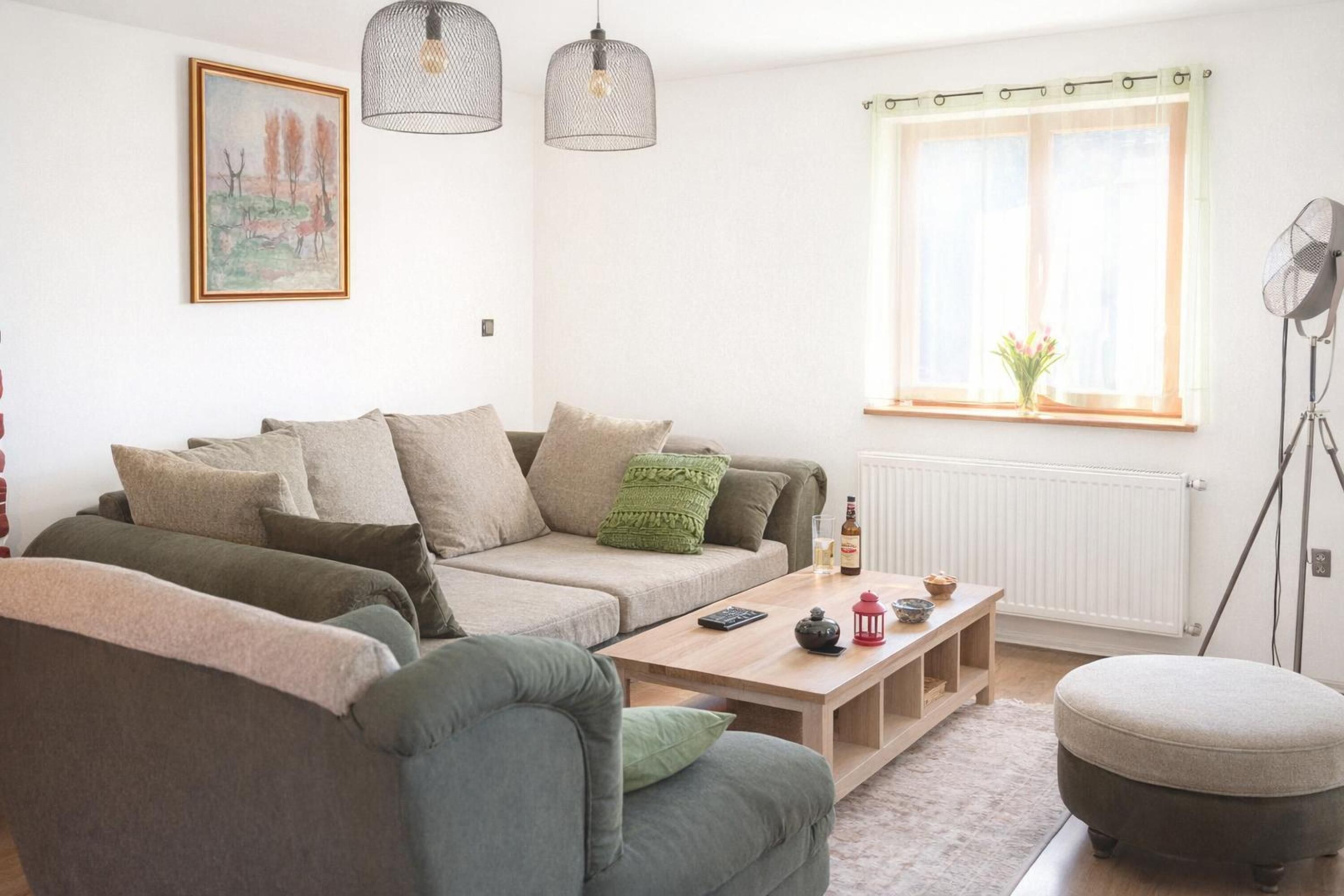 Una sala de estar moderna con un sofá, un reposapiés y una mesa de centro. Se ve una ventana con cortinas y un radiador.