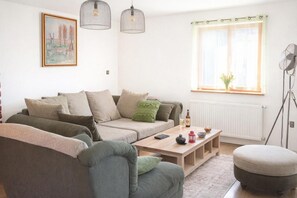 Ein modernes Wohnzimmer mit Sofa, Hocker und Couchtisch. Ein Fenster mit Vorhängen und eine Heizung sind ebenfalls vorhanden.