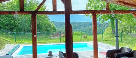 A covered outdoor area with a pool, wicker furniture, and a view of a forested landscape.