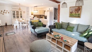 Una sala moderna con un sofá gris, una mesa de centro de madera y un reposapiés gris. Hay una zona de comedor con una mesa y sillas blancas, y una cocina al fondo.