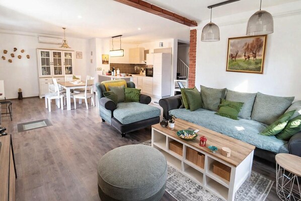 Ein modernes Wohnzimmer mit einem grauen Sofa, einem Holztisch und einem grauen Hocker. Im Hintergrund befindet sich ein Essbereich mit einem weißen Tisch und Stühlen sowie eine Küche.