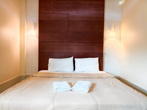 Una cama bien tendida con sábanas blancas y dos almohadas, un cabecero de madera y lámparas fijadas a la pared.