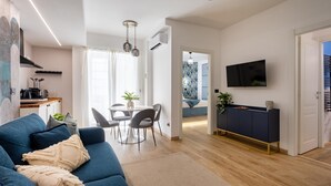 Un soggiorno moderno con un divano blu, un'area pranzo con tavolo e sedie, una zona cucina con macchina del caffè e una TV a parete.