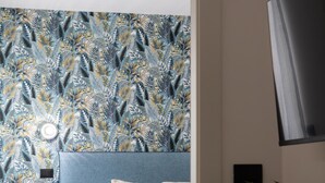 Una camera da letto con carta da parati decorata, un letto con lenzuola blu, un comodino, una pianta in vaso e un televisore montato a parete.