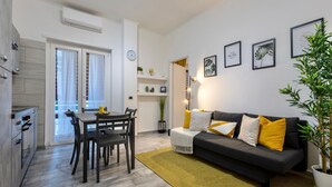 Una sala de estar moderna con sofá, mesa de comedor y sillas. Cuenta con una zona de cocina con armarios y una ventana.