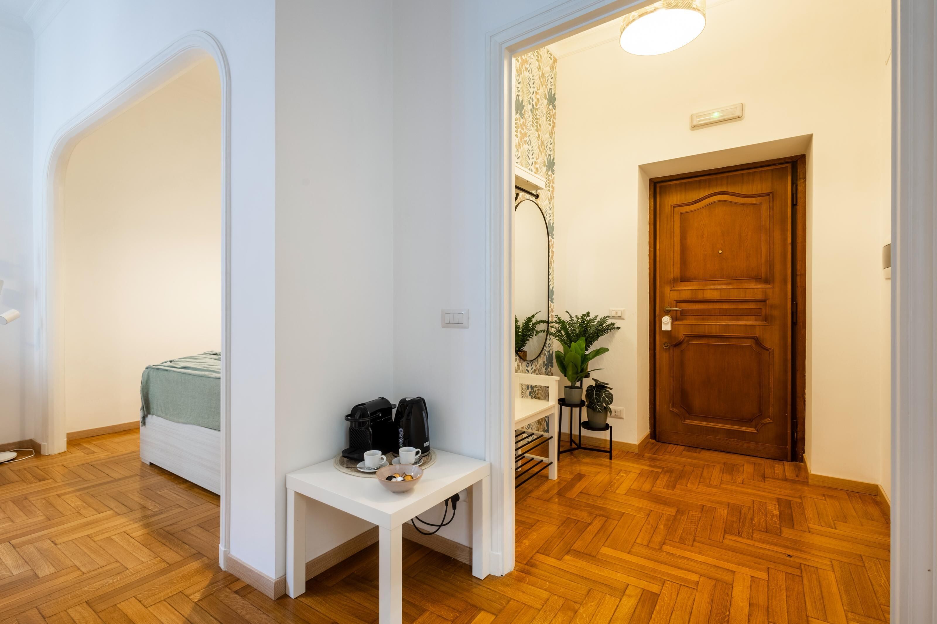 Ein Hotelzimmer mit Bett, kleinem Tisch mit Kaffeemaschine, Pflanze und Holztür.