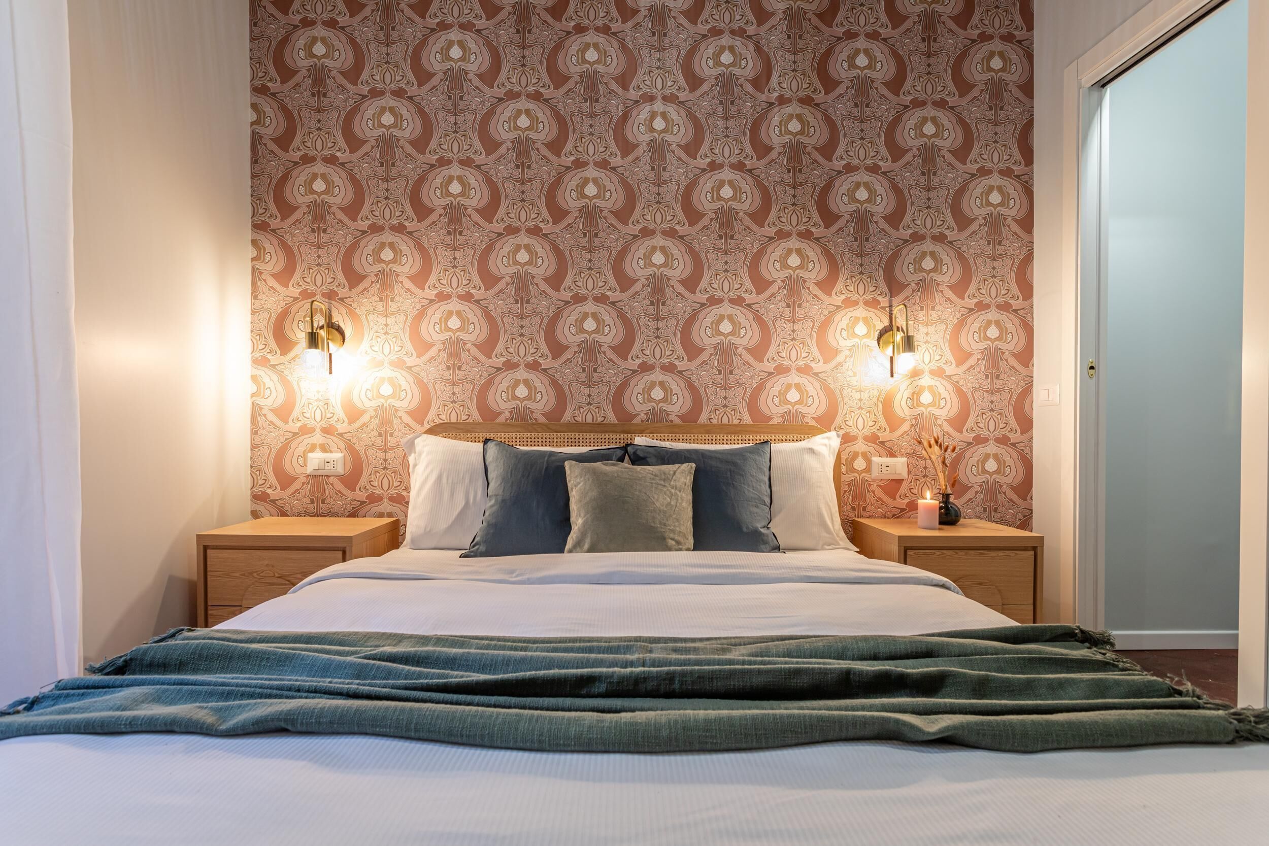 A neatly made bed with a green blanket, two bedside tables with lamps, and a patterned wallpaper.