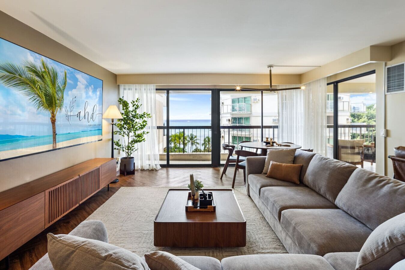 A spacious living room with a large sofa, a coffee table, and a painting of a beach scene. There is a balcony with a view of the ocean and palm trees.