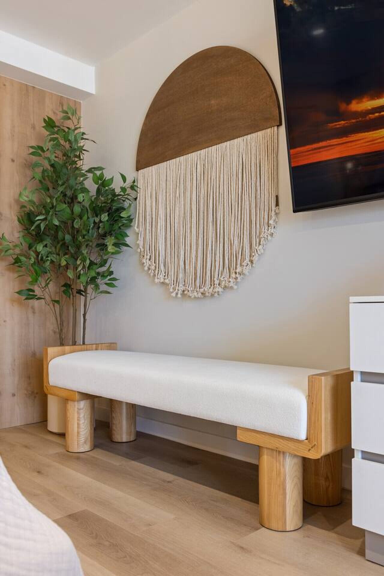 Een moderne slaapkamer met een houten bankje, een macramé wanddecoratie, een potplant en een schilderij.