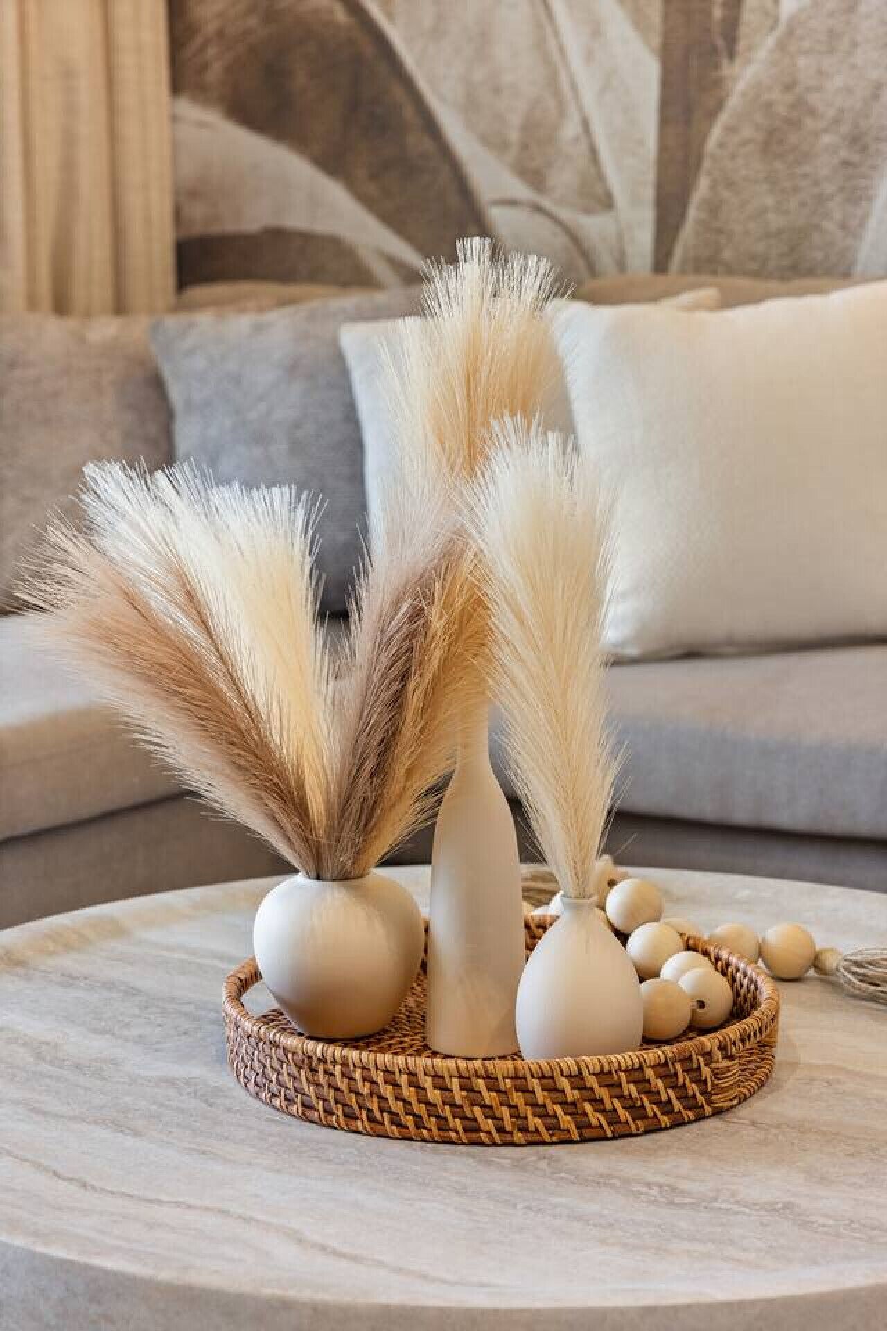 A decorative arrangement of white vases and pampas grass on a round marble table, placed on a woven basket.