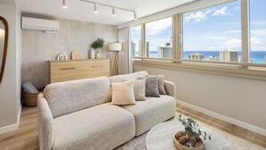 A modern living room with a beige sofa, a wooden side table, and a view of the city skyline through large windows. - Waikiki Ocean Luxe Retreat High Floor Suite (Honolulu)