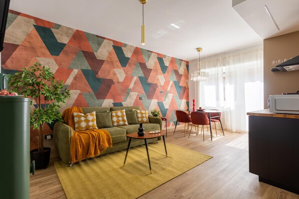 Een moderne woonkamer met een wand met een geometrisch patroon, een bank met kussens, een salontafel, een eethoek met stoelen en een keuken met een magnetron.