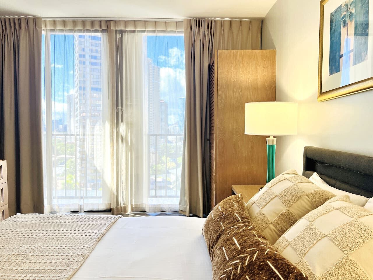 Habitación de hotel con una cama grande, una mesita de noche con lámpara y vistas a edificios altos a través de las ventanas.