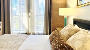 Een hotelkamer met een groot bed, een nachtkastje met een lamp en uitzicht op hoge gebouwen door de ramen.