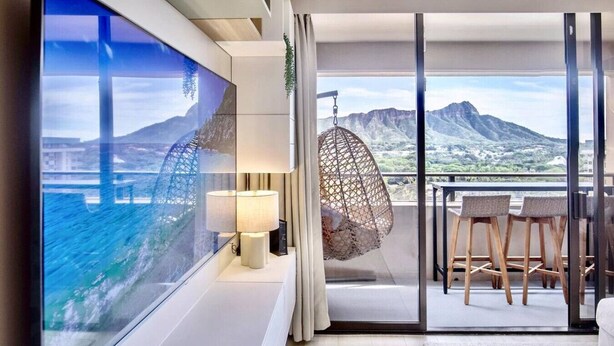 Ein modernes Zimmer mit großem Fernseher, der eine reizvolle Aussicht zeigt, einem Hängesessel, einem Tisch mit Stühlen und einem Balkon mit Blick auf Berge und eine Brücke.