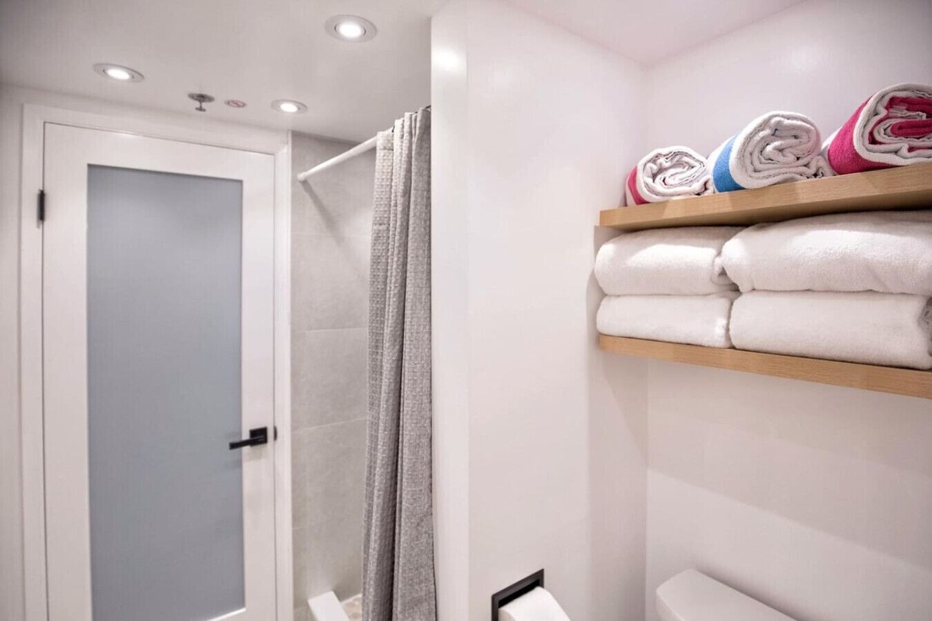 A bathroom with a glass shower enclosure, a white toilet, and a shelf with folded towels.