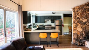 A modern kitchen with dark cabinets, a central island with two orange chairs, and a stone wall feature. - Hot Tub, Walk to Town, Lake Access, Fireplace, Fenced Yard (Twain Harte)